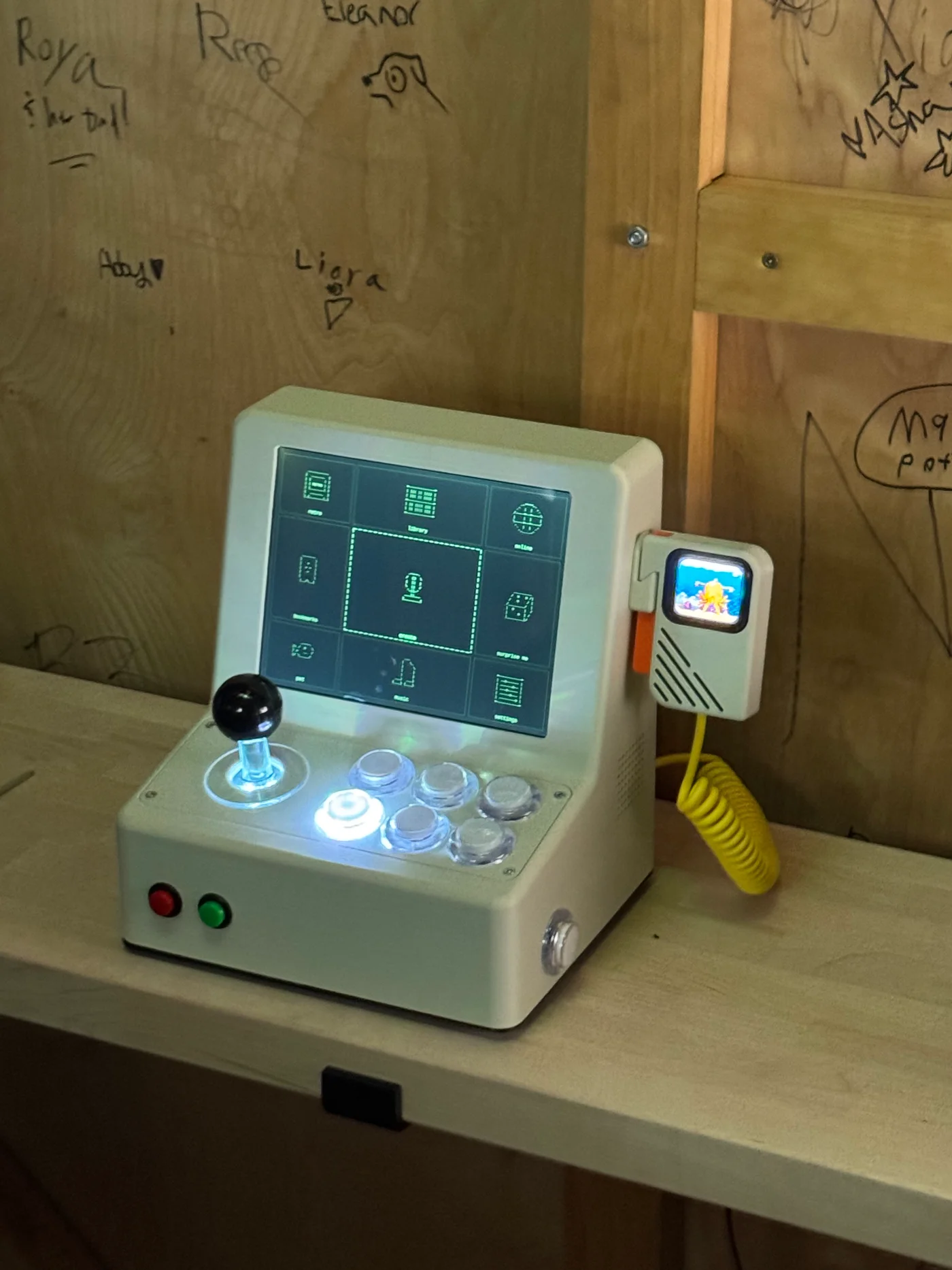 VibeConsole on a wooden shelf, with illuminated arcade buttons and a handheld companion connected by a yellow coiled cable.